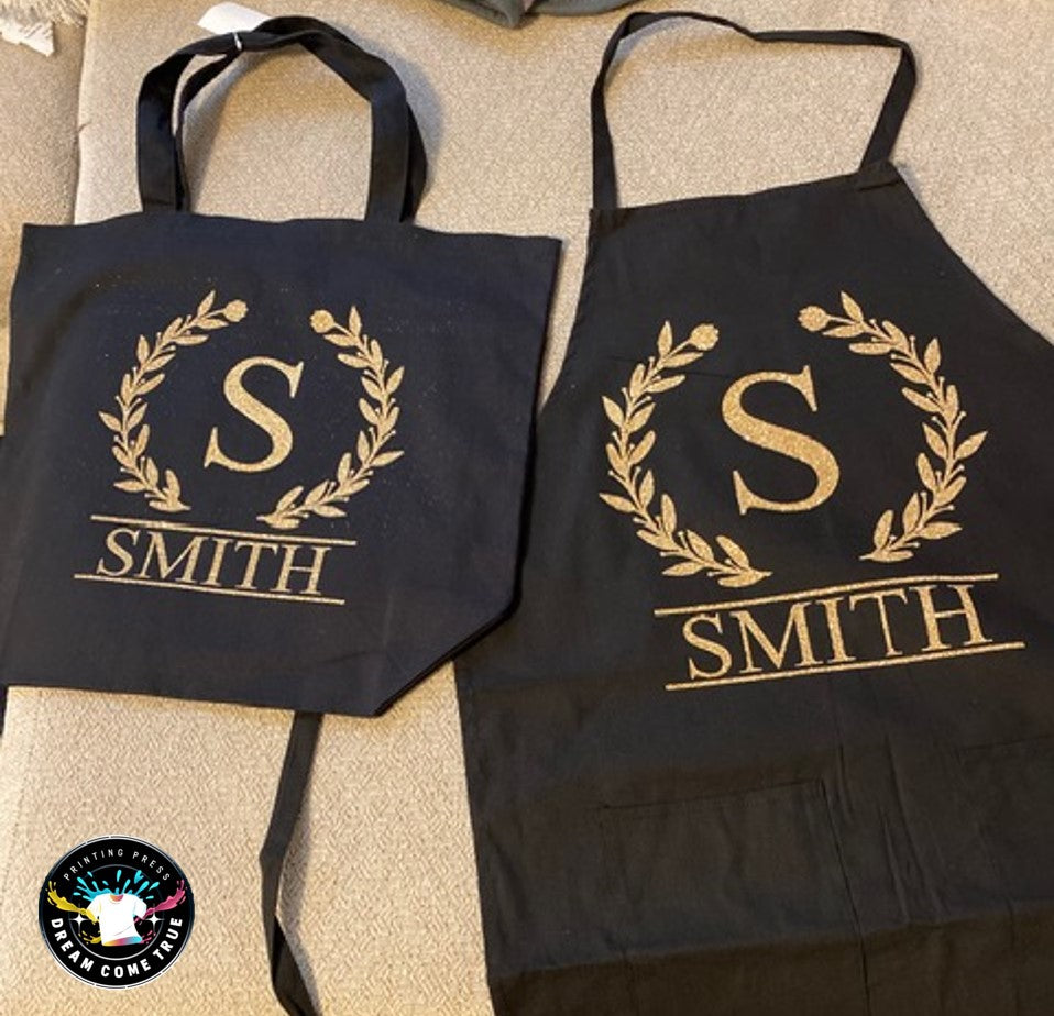 Black tote bag and apron with 'S Smith' design on a beige surface at D.C.T Printing Press.