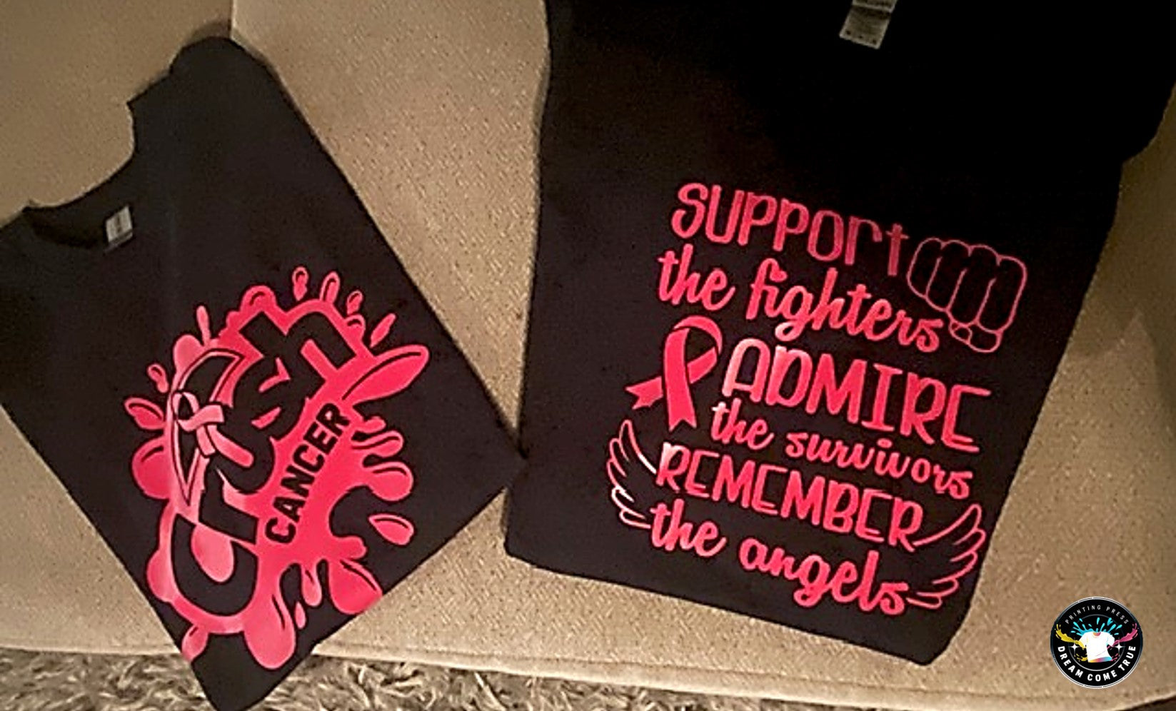 Two black shirts with pink cancer awareness designs on a beige surface at D.C.T. Printing Press.