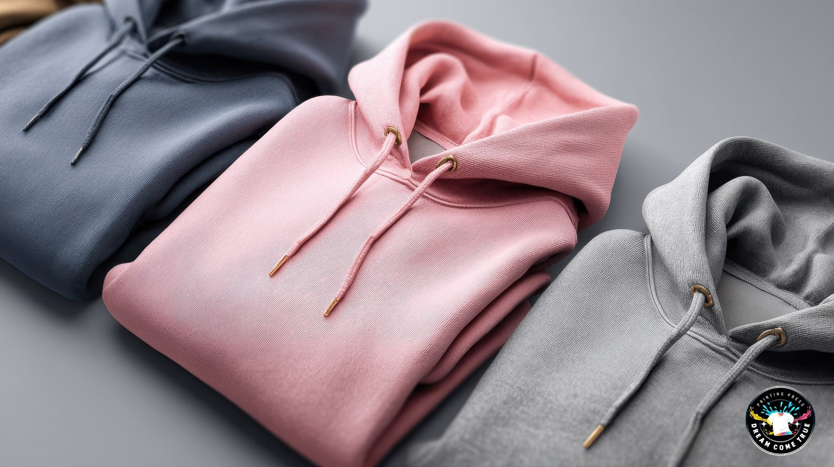 Three folded hoodies in gray, pink, and gray on a flat surface, customize at Dream Come True Printing Press.